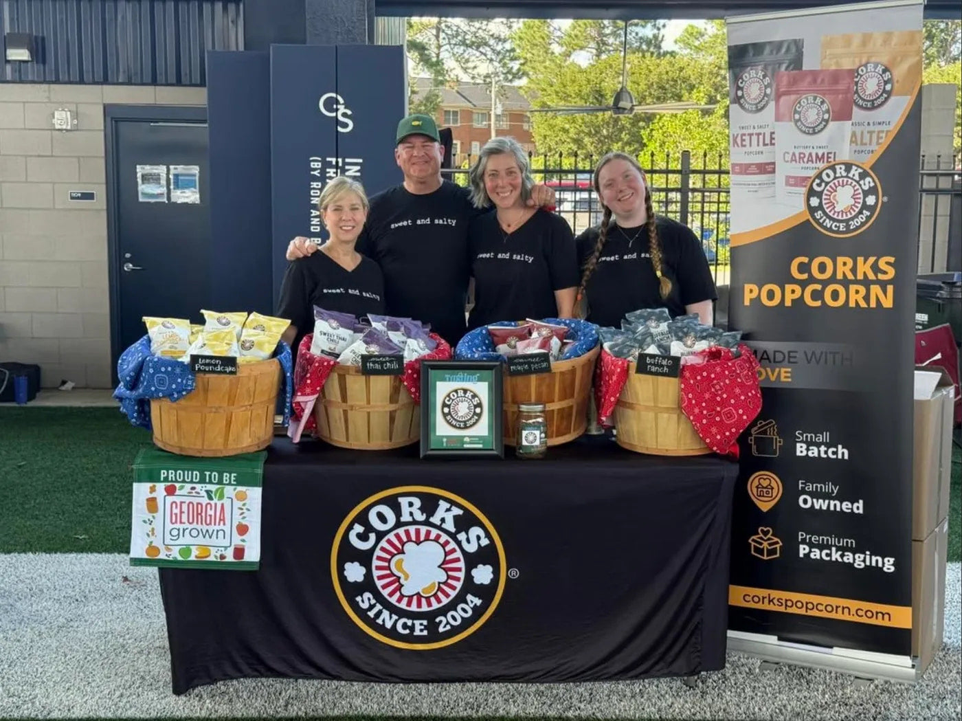 The Corks Popcorn team at their booth with baskets of popcorn and a Corks Popcorn banner